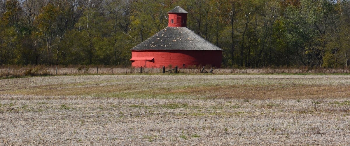 A red barn with a conical roof surrounded by a field and trees.