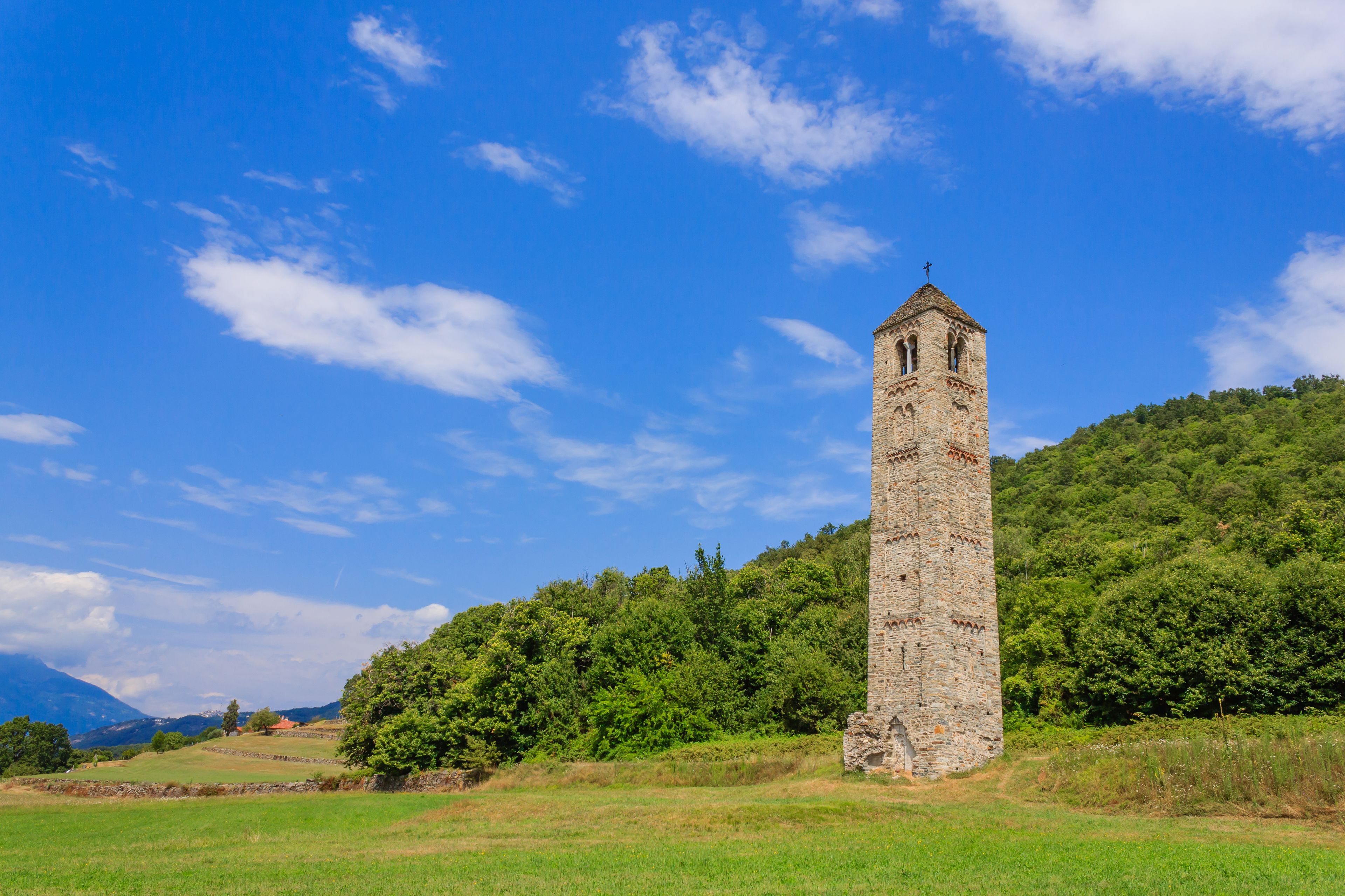 Ein hoher, steinerner Turm mit einem Kreuz auf der Spitze, umgeben von üppigen grünen Hügeln und wolkenlosem, blauem Himmel.