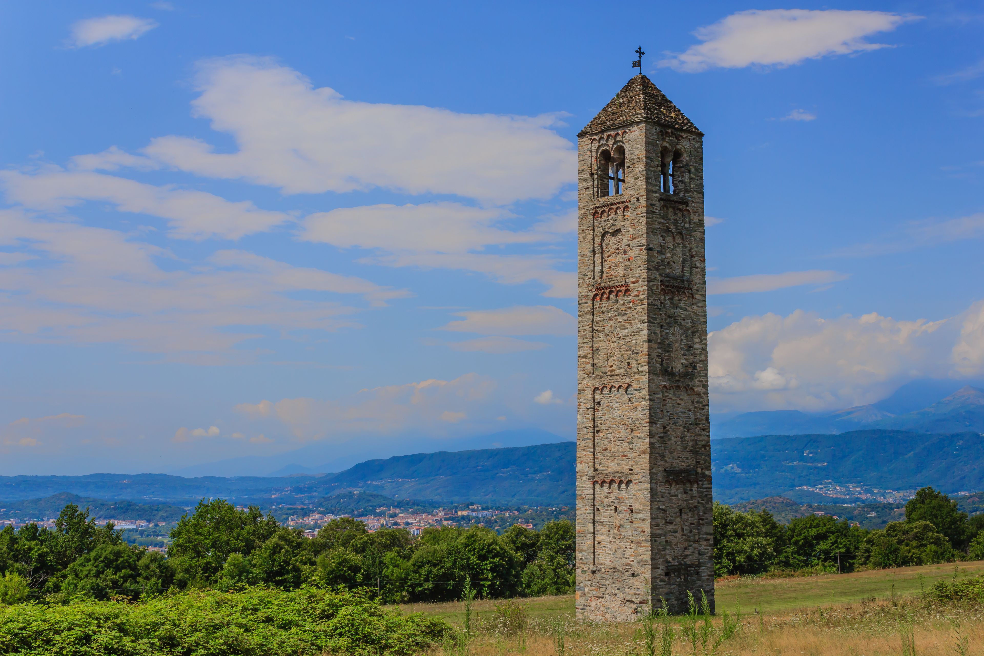 Ein hoher, steinerner Turm mit Kreuz, umgeben von Grün und einer bergigen Landschaft im Hintergrund.