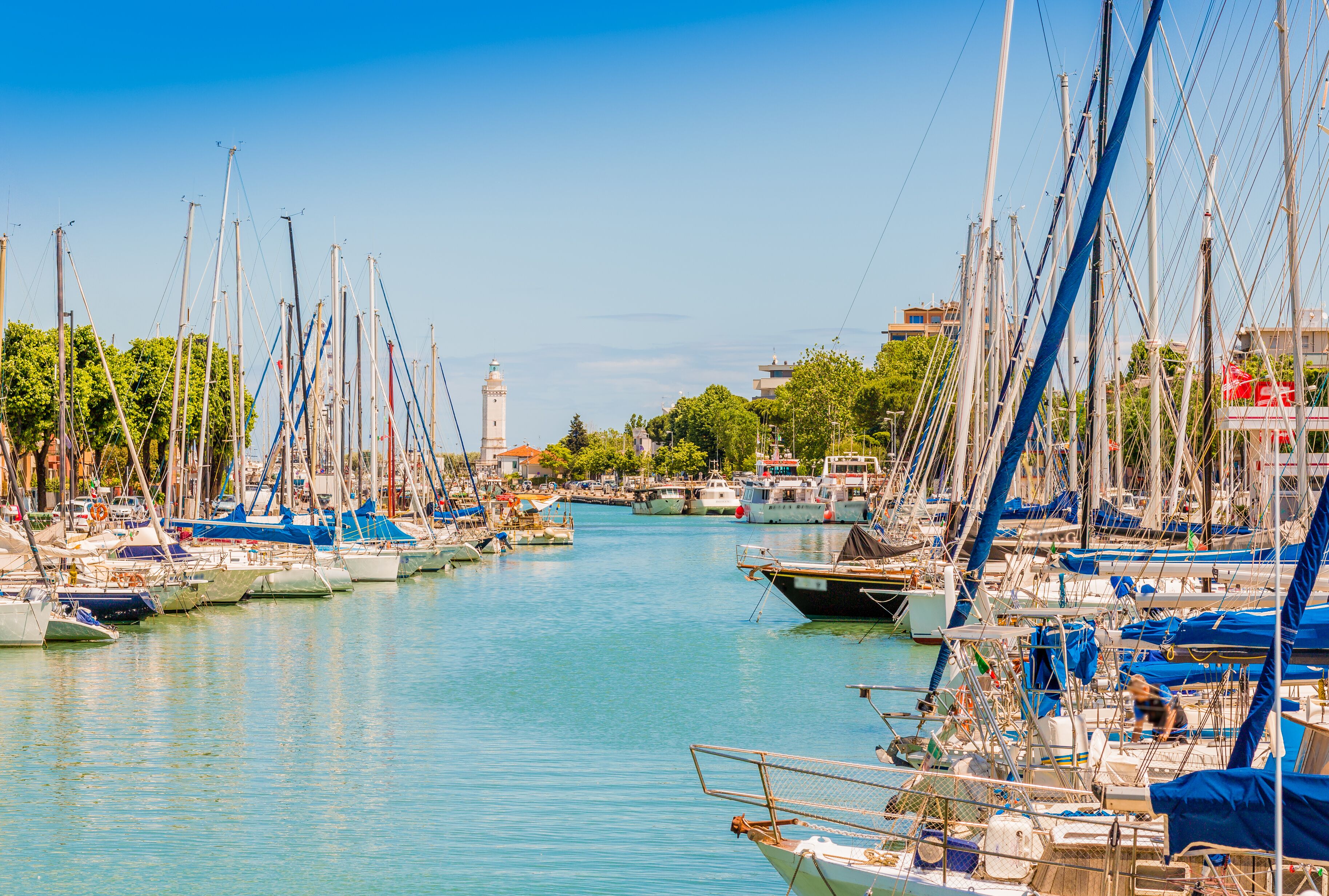A marina with numerous sailboats docked along a calm waterway, surrounded by greenery and buildings.