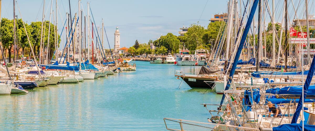 A marina with numerous sailboats docked along a calm waterway, surrounded by greenery and buildings.