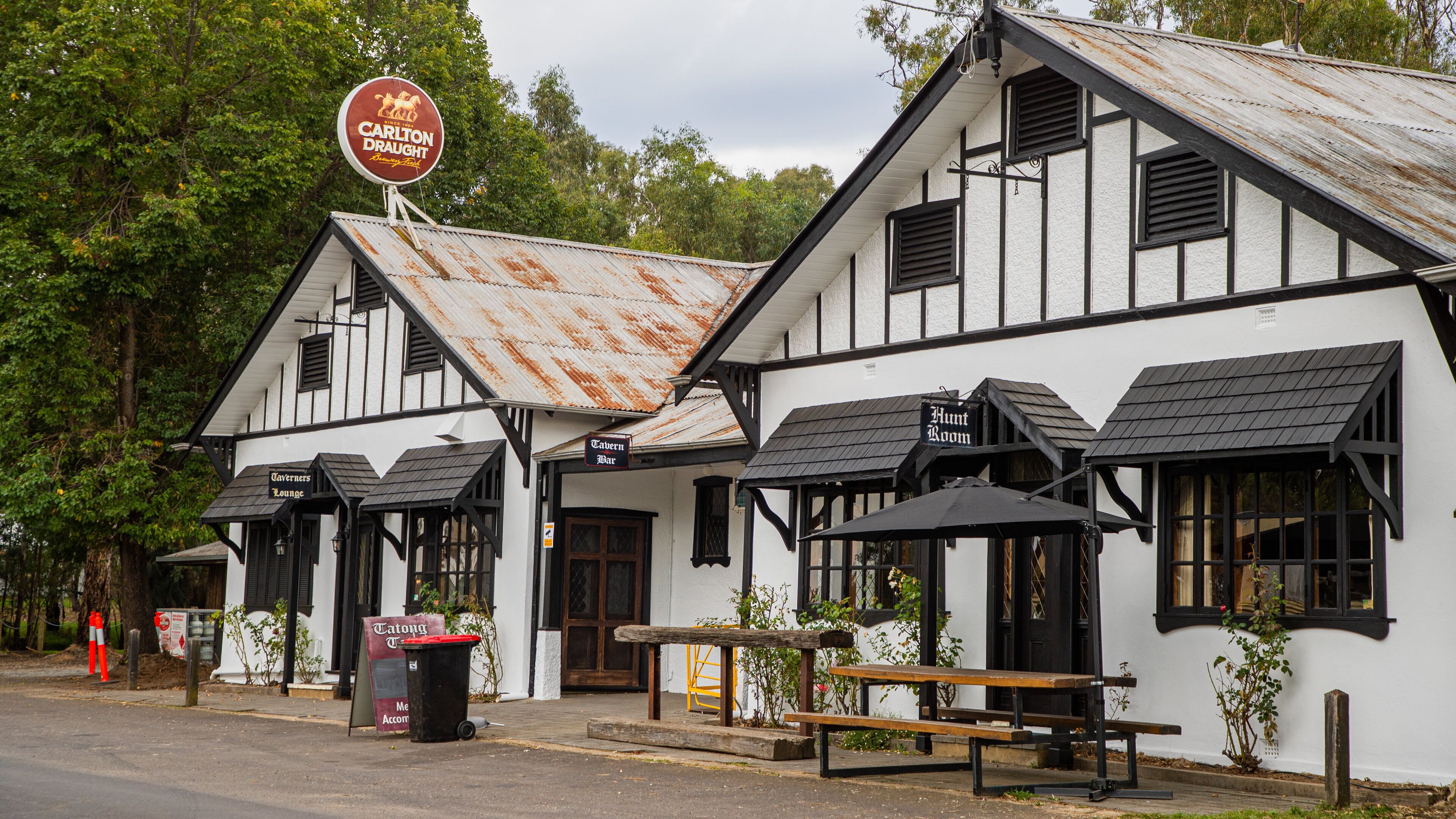 A white building with black trim and a rusty roof. There is a sign that says "Carlton Draught" and a sign that says "Hunt Room".
