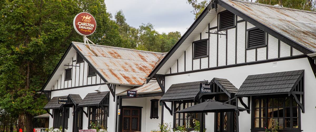 A white building with black trim and a rusty roof. There is a sign that says "Carlton Draught" and a sign that says "Hunt Room".
