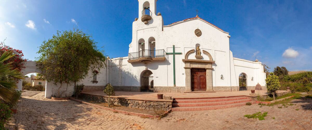 A white church with a bell tower and a cross, surrounded by greenery and cobblestone paths.