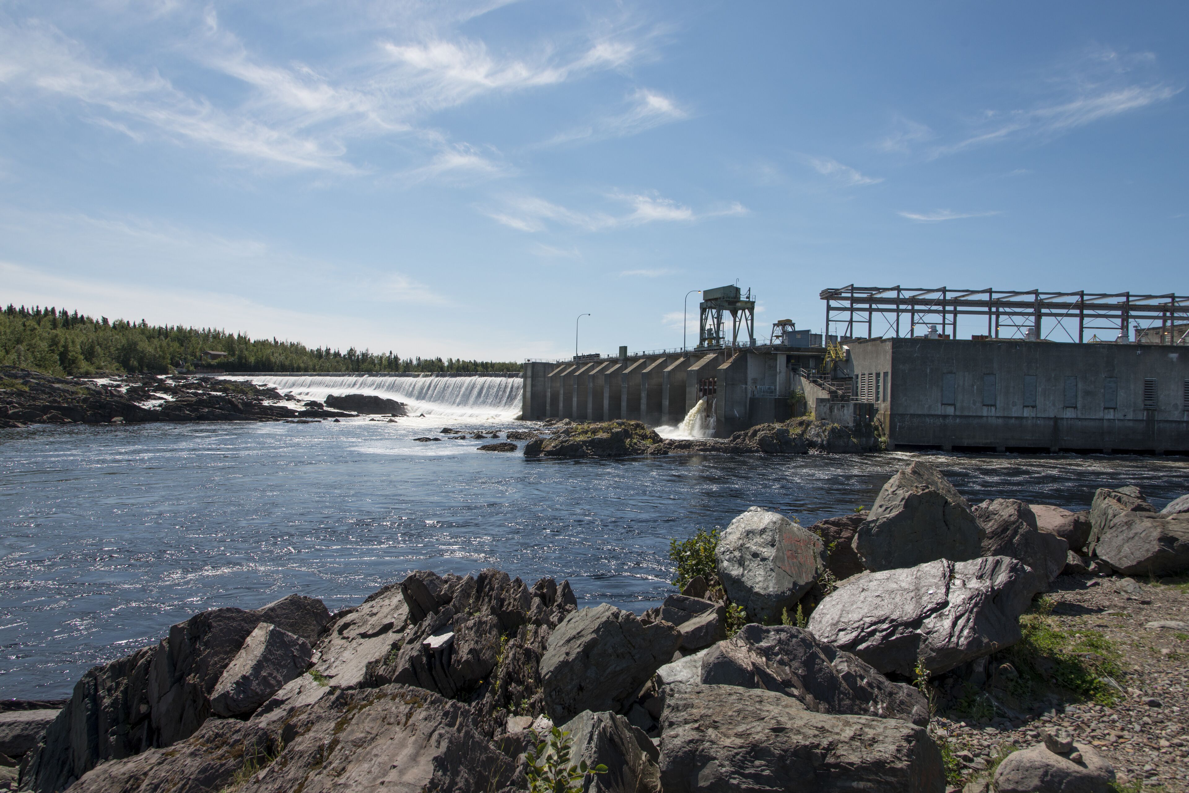 A dam with water flowing over it, a rocky shoreline, and a forest in the background.