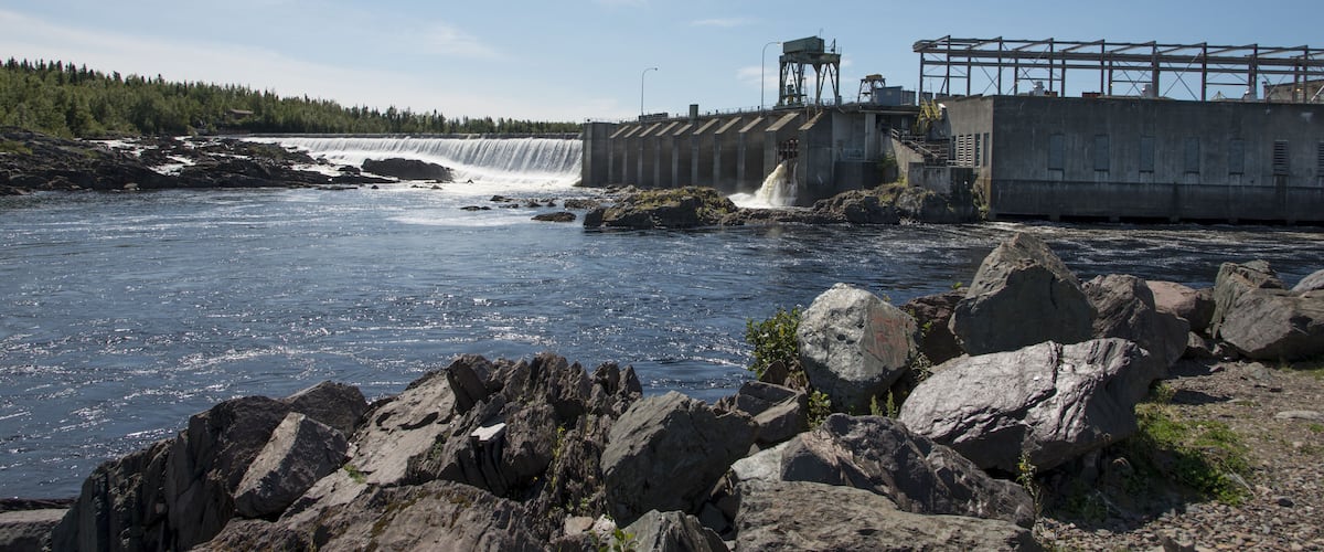 A dam with water flowing over it, a rocky shoreline, and a forest in the background.