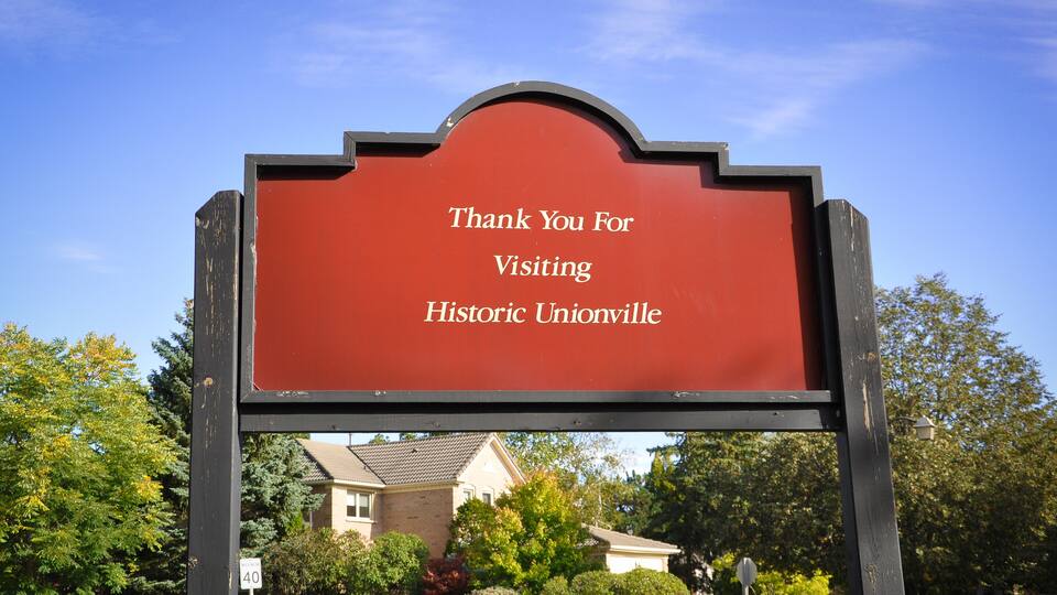 A red sign with white text that reads "Thank You For Visiting Historic Unionville" set against a backdrop of a residential neighborhood with houses and trees.