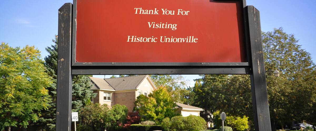 A red sign with white text that reads "Thank You For Visiting Historic Unionville" set against a backdrop of a residential neighborhood with houses and trees.