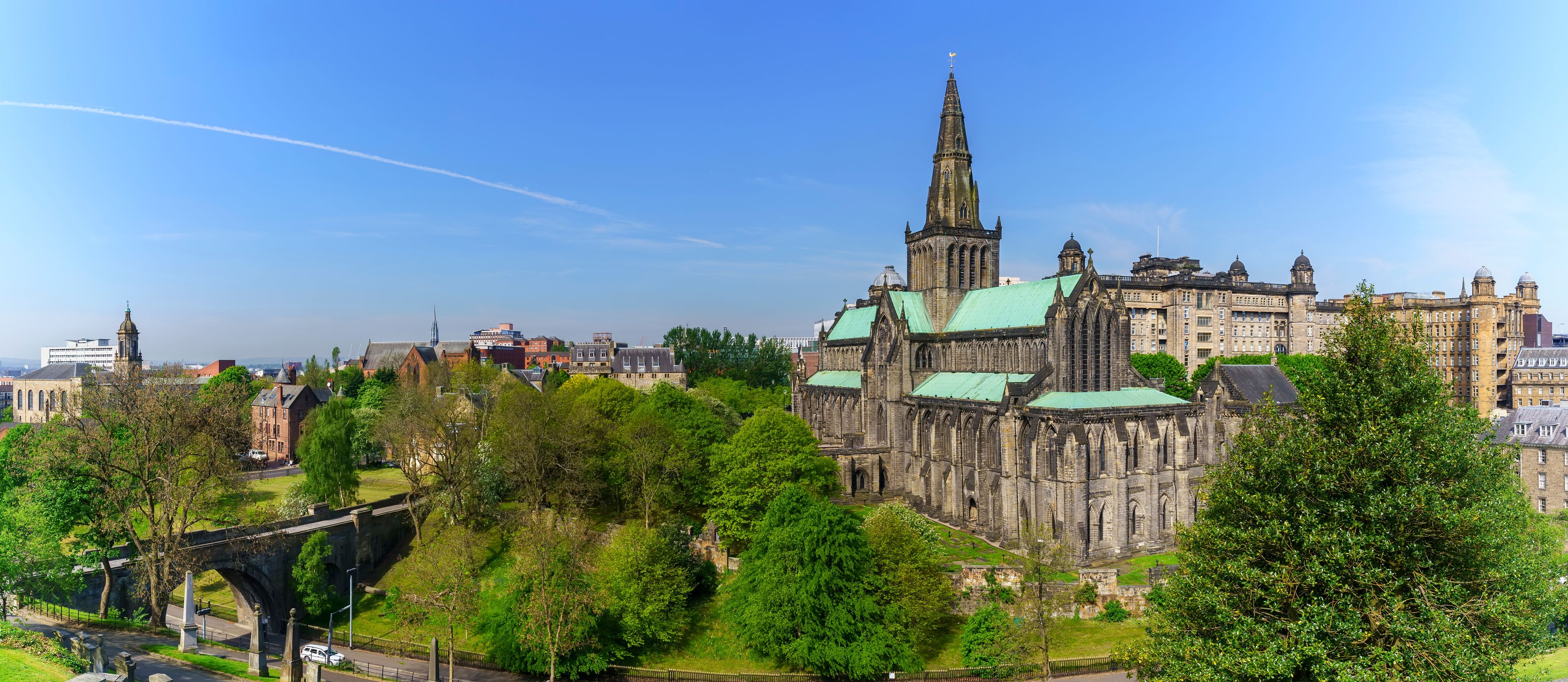 Una catedral histórica con una aguja alta, rodeada de exuberante vegetación y edificios modernos.