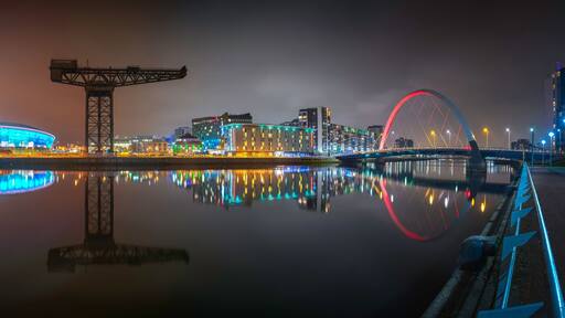 A cityscape at night with illuminated buildings, a bridge, and a calm river reflecting the lights.