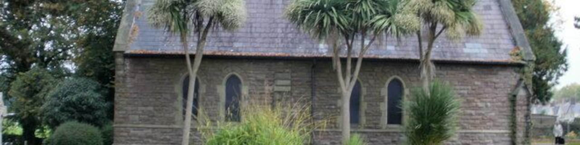 Igreja de pedra com telhado inclinado, rodeada por um cemitério com lápides e palmeiras.