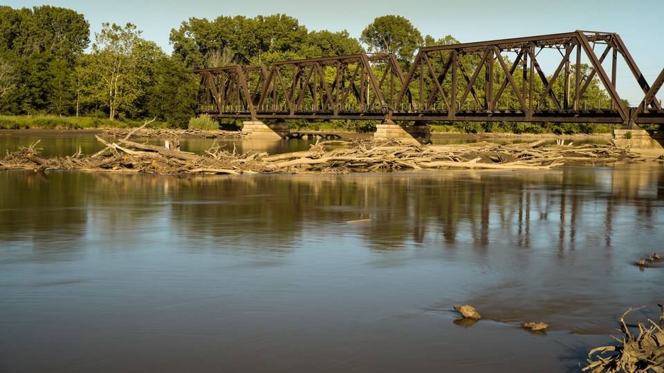 A rusted metal bridge over a calm river with trees in the background.