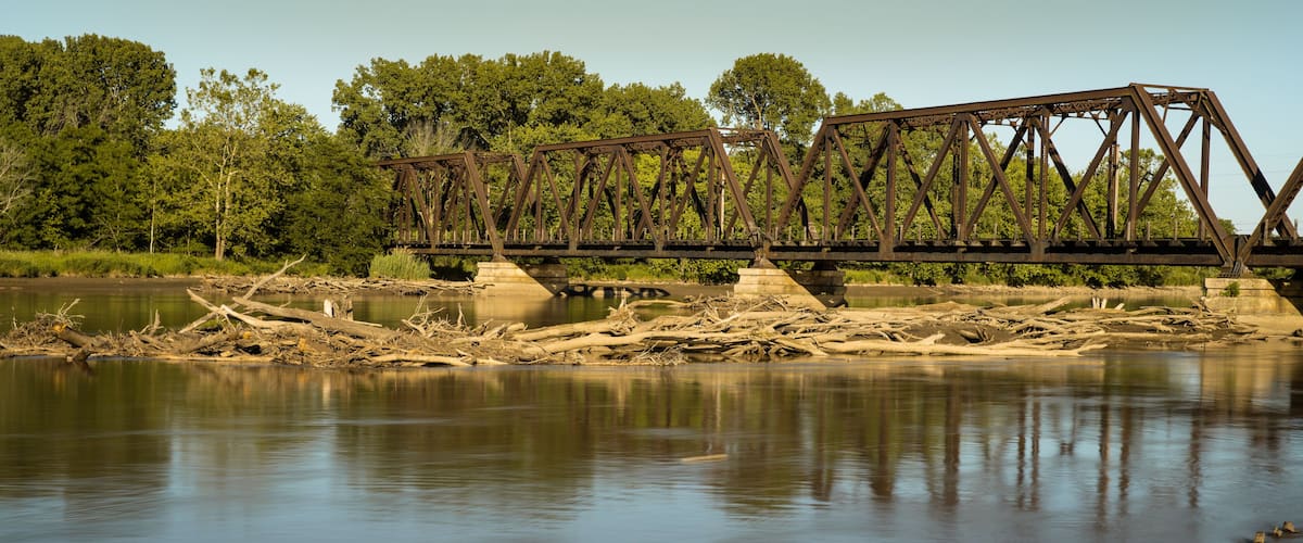 A rusted metal bridge over a calm river with trees in the background.