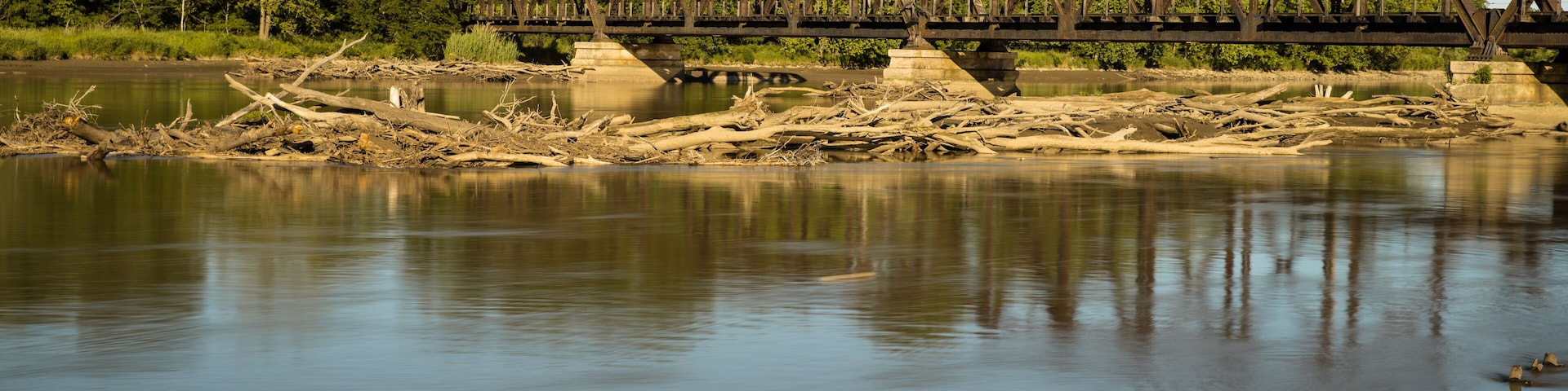 A rusted metal bridge over a calm river with trees in the background.