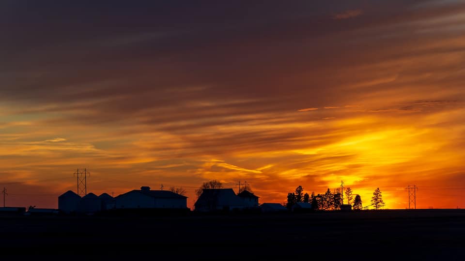 A sunset over a rural landscape with houses and power lines silhouetted against a vibrant sky.