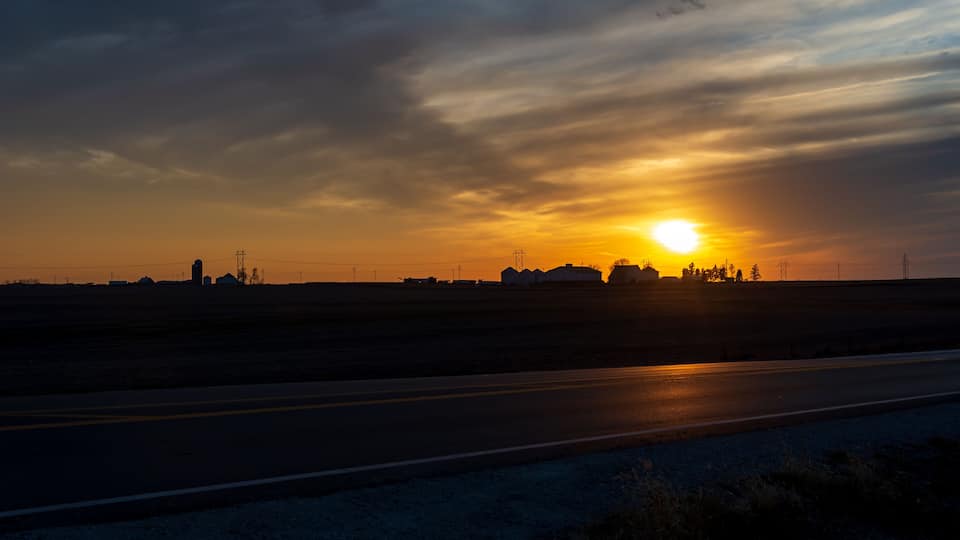 A sunset over a flat landscape with a road and distant buildings.
