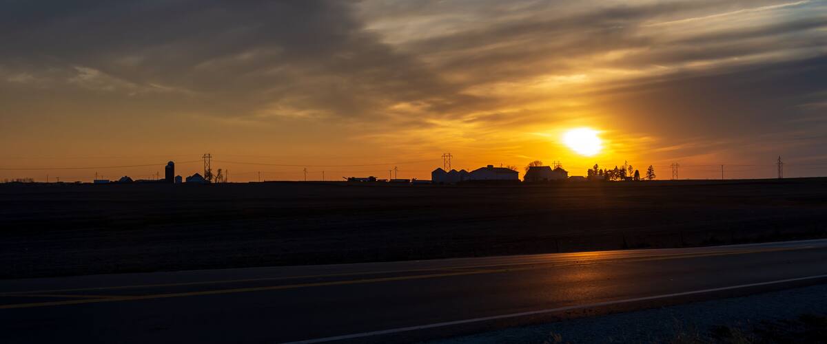 A sunset over a flat landscape with a road and distant buildings.