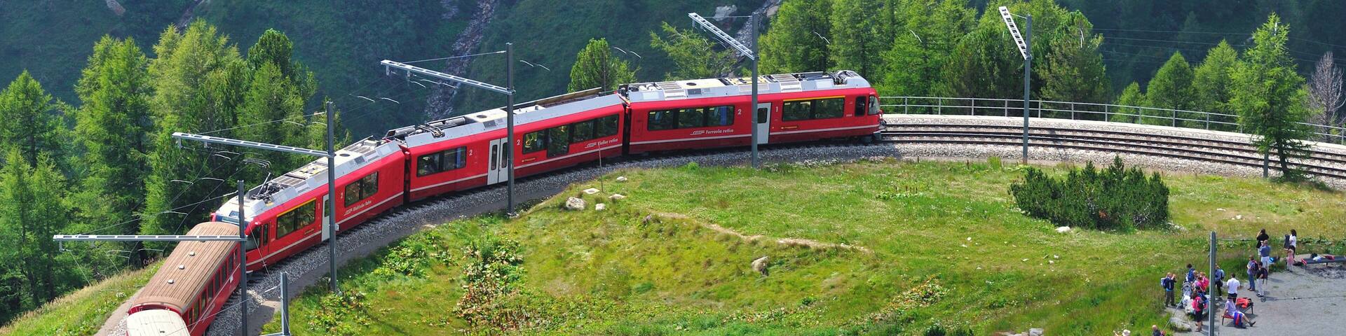 A red train traveling through a mountainous area with a small lake and people standing by the shore.