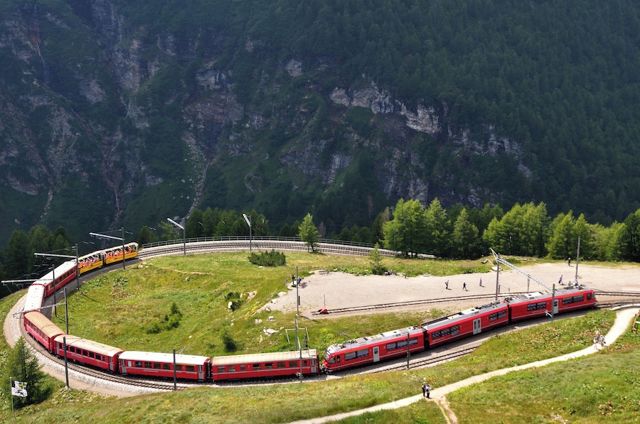 Un tren rojo sobre una vía curva que atraviesa una zona montañosa con árboles verdes y riscos rocosos.