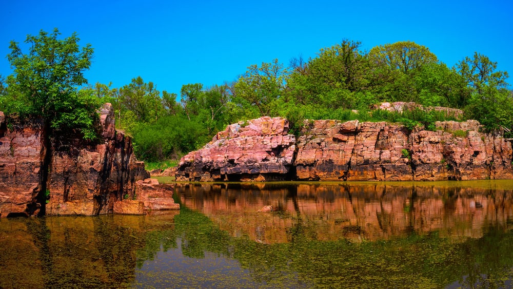 A calm lake with clear reflections, surrounded by rocky cliffs and lush green trees.