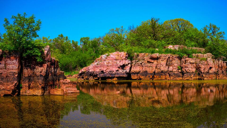 A calm lake with clear reflections, surrounded by rocky cliffs and lush green trees.