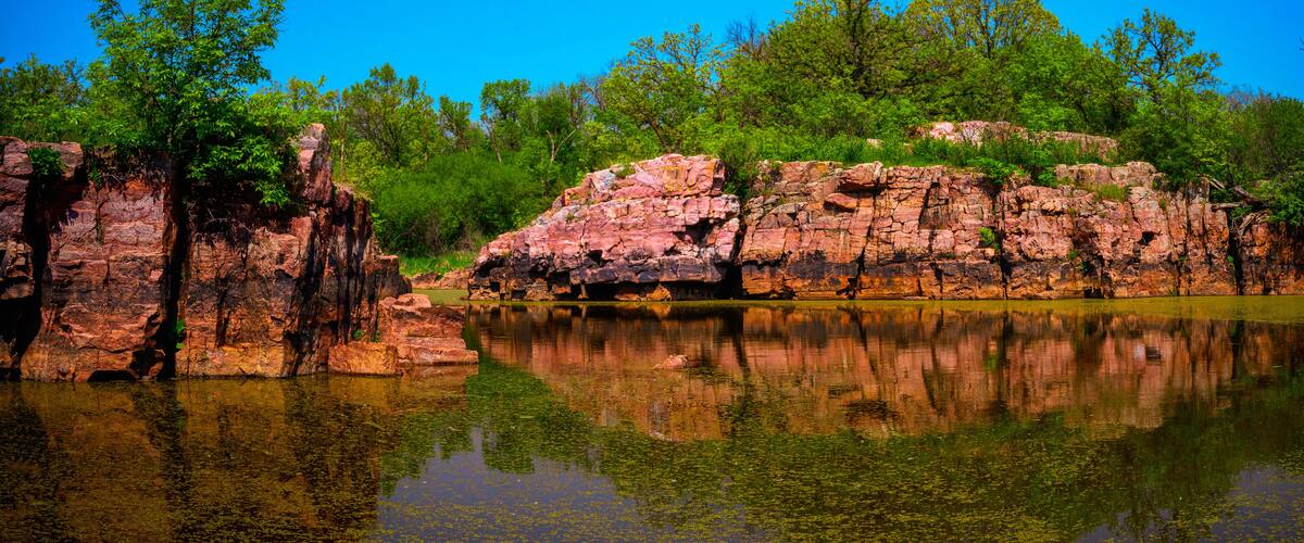 A calm lake with clear reflections, surrounded by rocky cliffs and lush green trees.