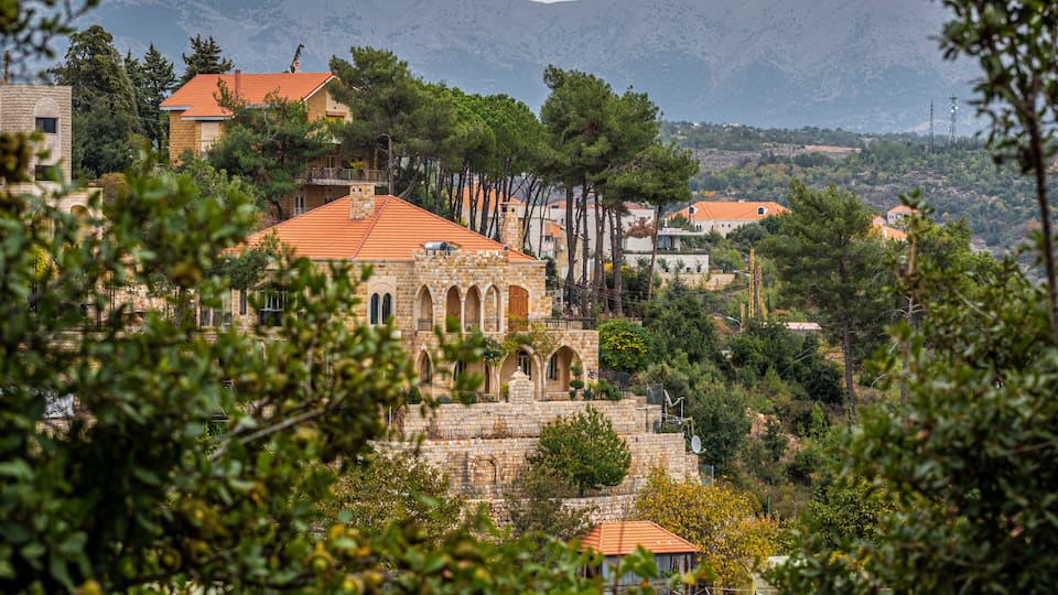 A stone building with a red roof, surrounded by trees and hills in the background.