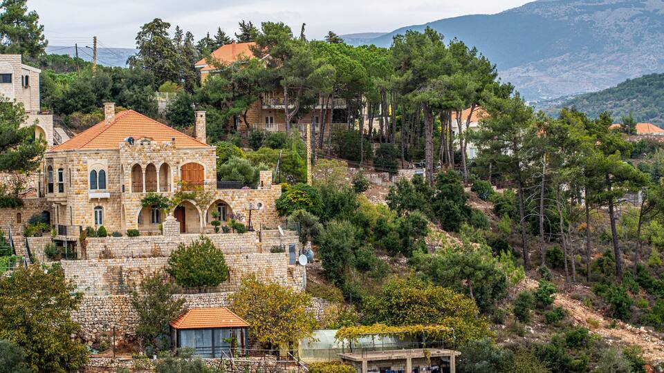 A stone building with a red-tiled roof, surrounded by trees and hills.