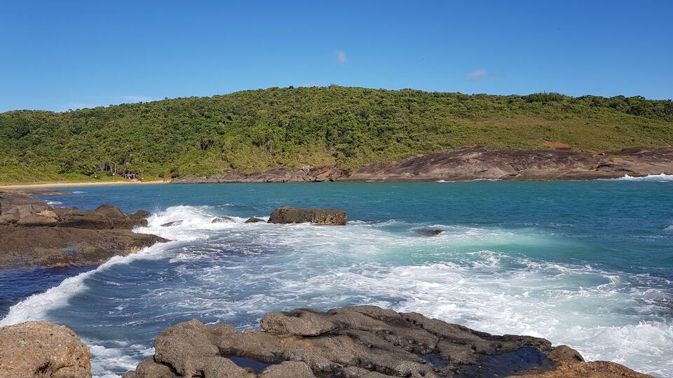 A coastal landscape with rocky shores, clear blue waters, and a forested hill in the background.