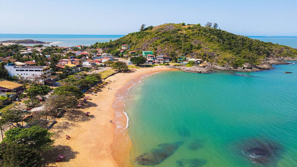 A coastal town with a sandy beach, clear turquoise waters, and a hill with buildings.
