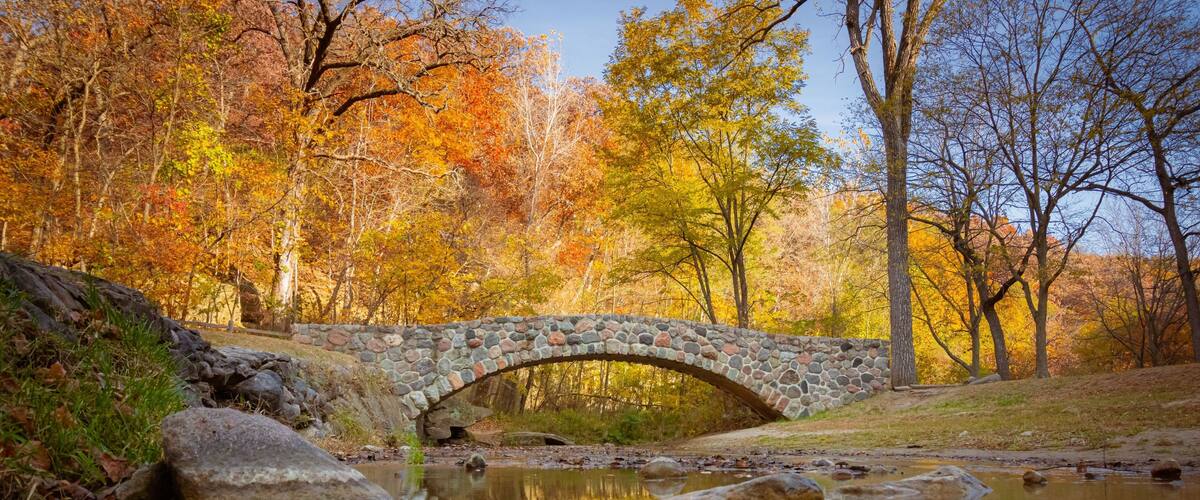 A stone bridge over a calm stream surrounded by autumn-colored trees.