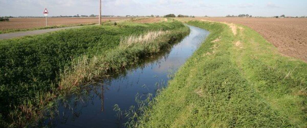 A narrow water channel with grassy banks, a dirt path, and a warning sign in a rural landscape.