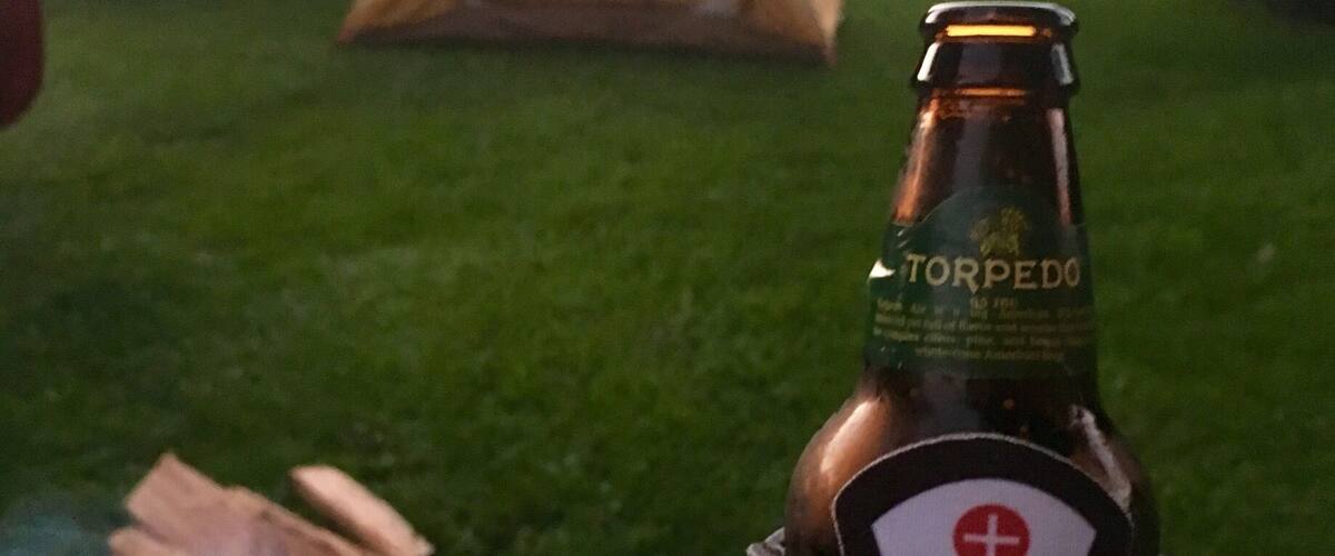 A hand holding a bottle of 'Courses Rock' beer with a tent and a campfire in the background.
