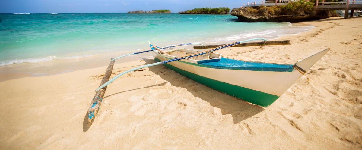 A beach with a boat, clear turquoise water, and a small hut on a rocky outcrop.