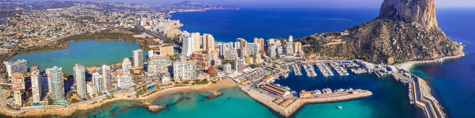A coastal cityscape with high-rise buildings, a marina, and a prominent rocky cliff.