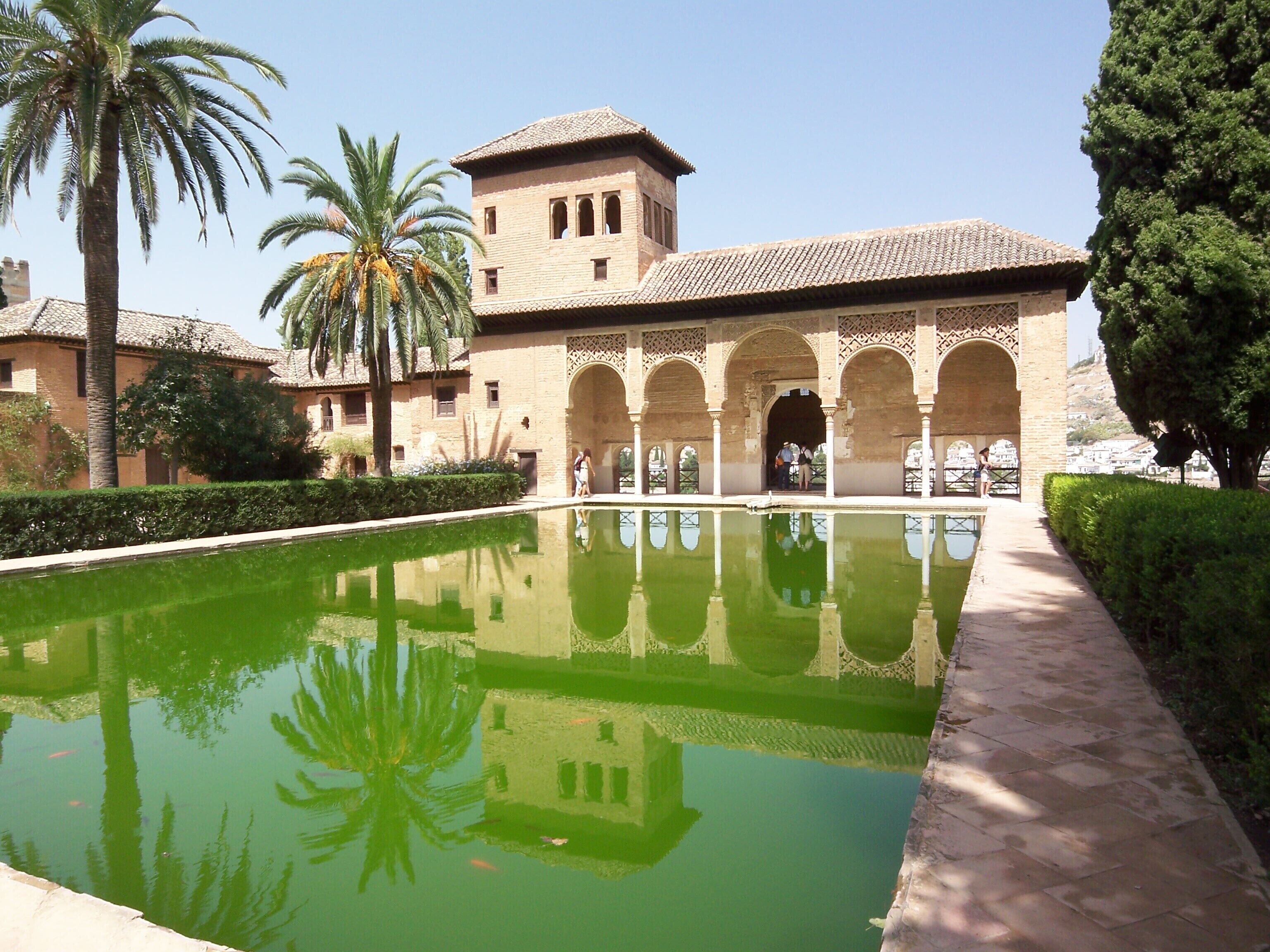 A courtyard with a pool, palm trees, and a building with arches and a tower.