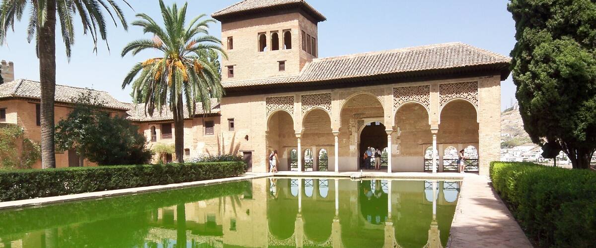 A courtyard with a pool, palm trees, and a building with arches and a tower.