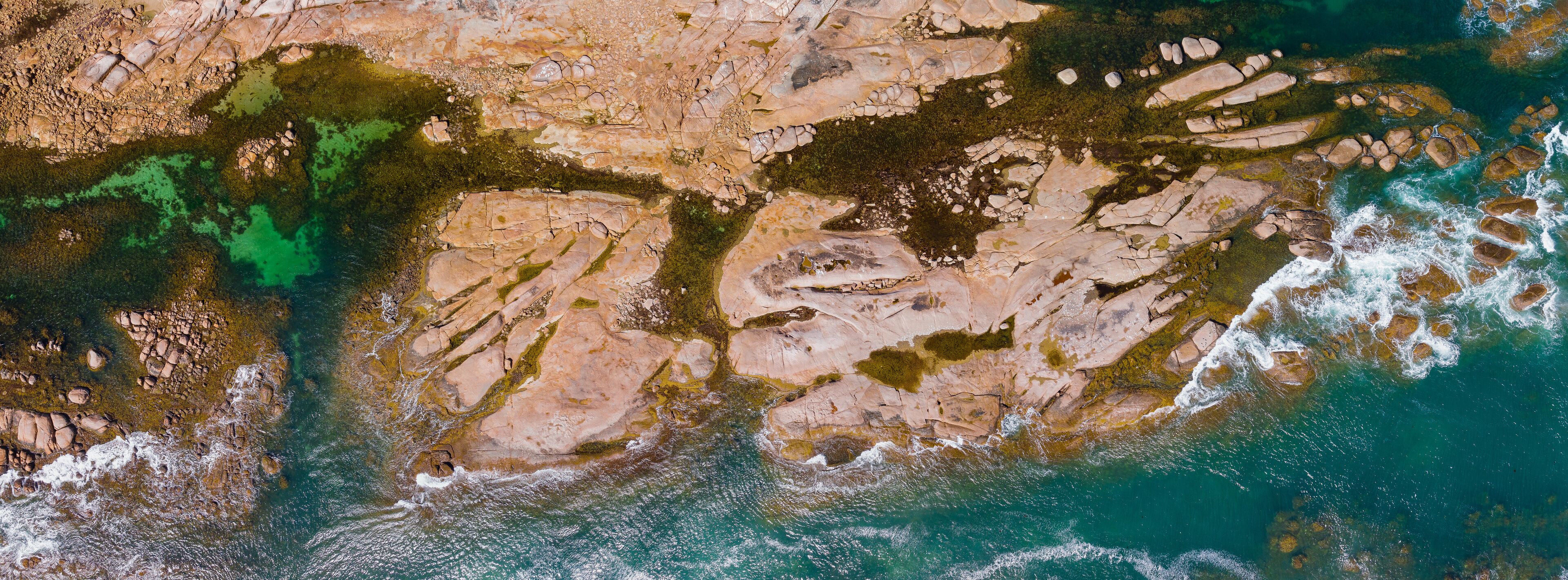 Aerial panorama view of water flowing around large granite ledges covered in lichen and weed
