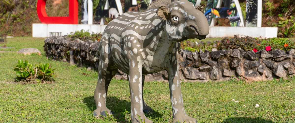 A sculpture of a stylized animal in a park with a backdrop of a large, colorful sign.