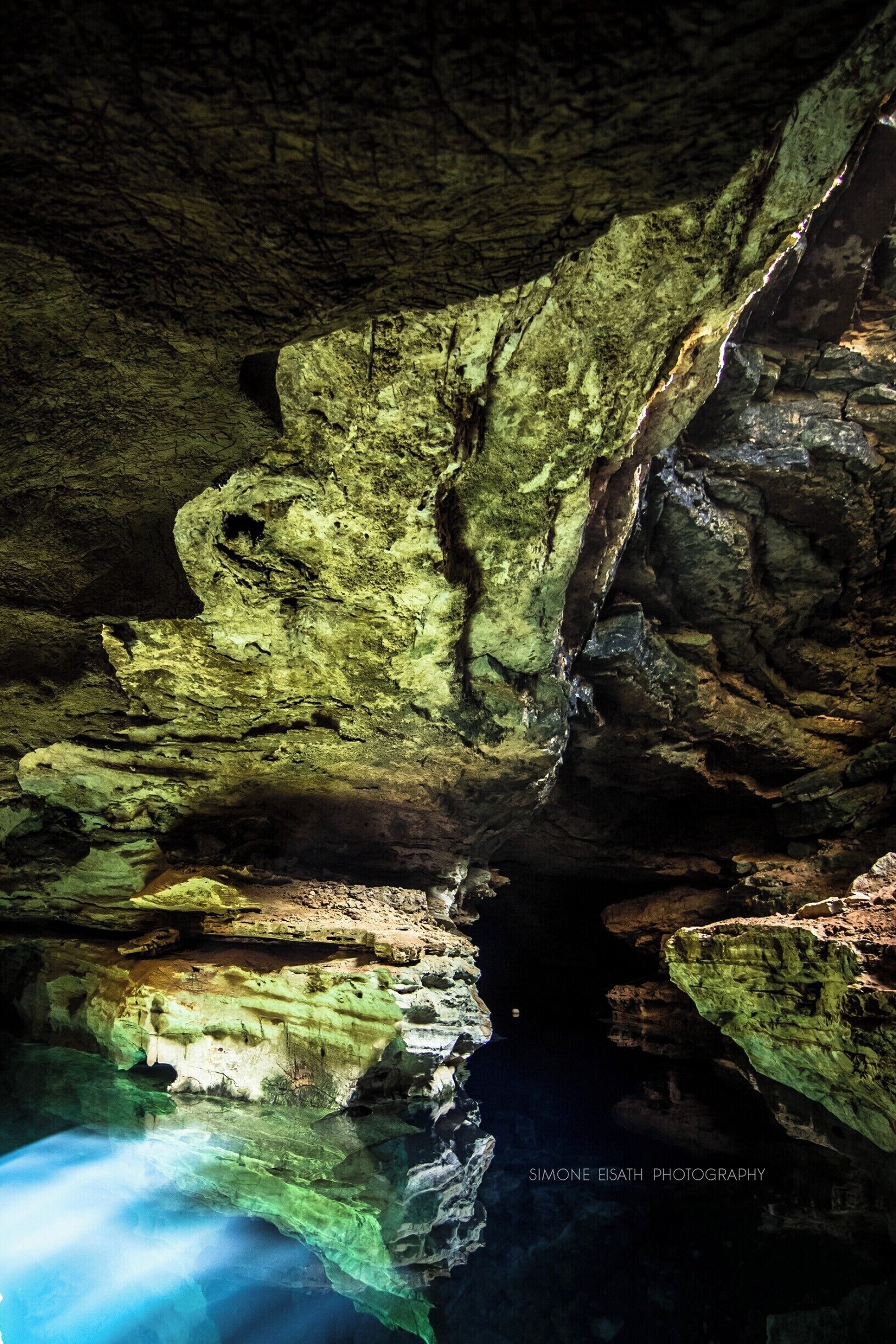 A cave with greenish rock formations and a clear water pool at the bottom.