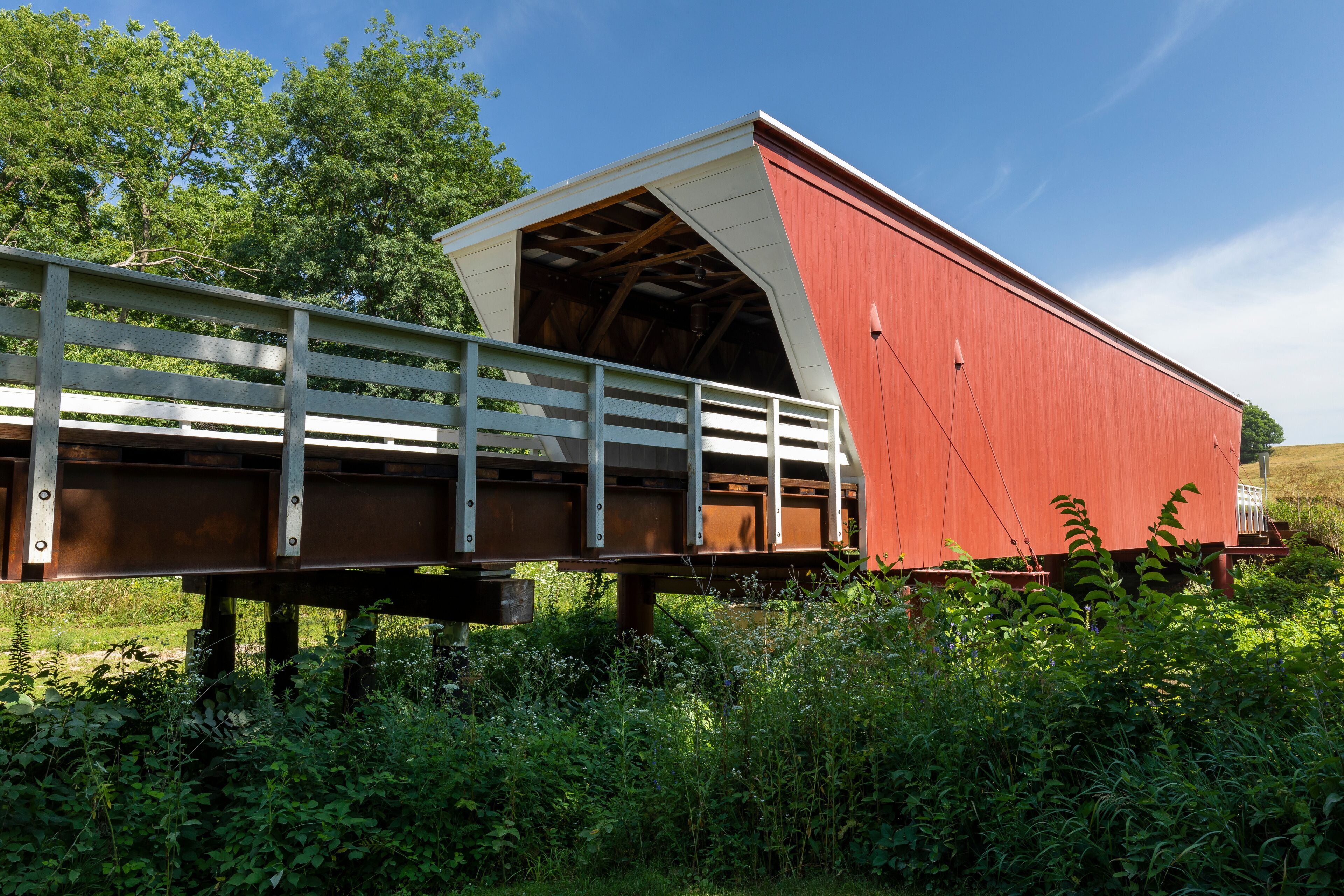 A covered bridge with a red roof and white walls, supported by wooden beams, crossing a grassy area with dense vegetation.