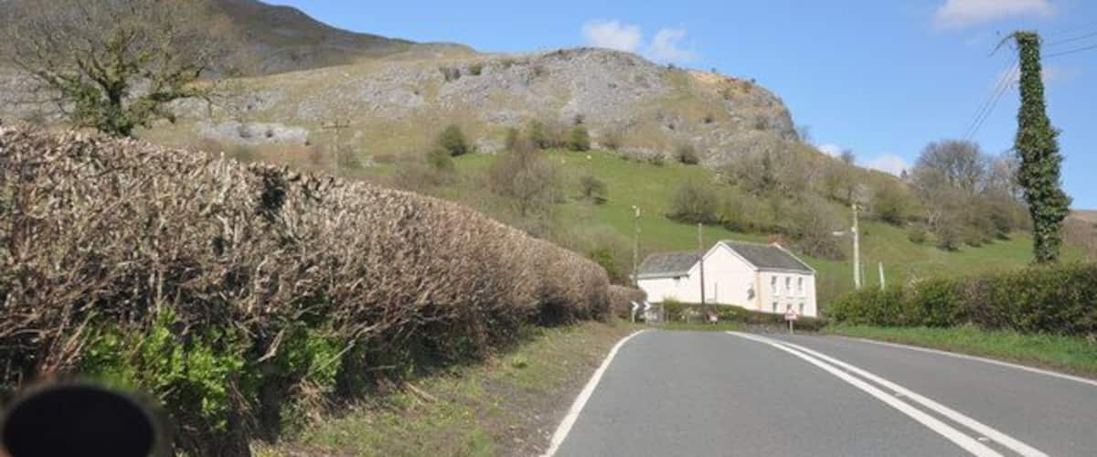 A rural road with a stone wall, a white house, and a hill in the background.