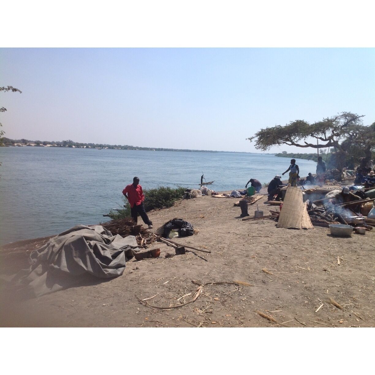 A riverside scene with people near a makeshift shelter, a boat, and a distant shoreline.