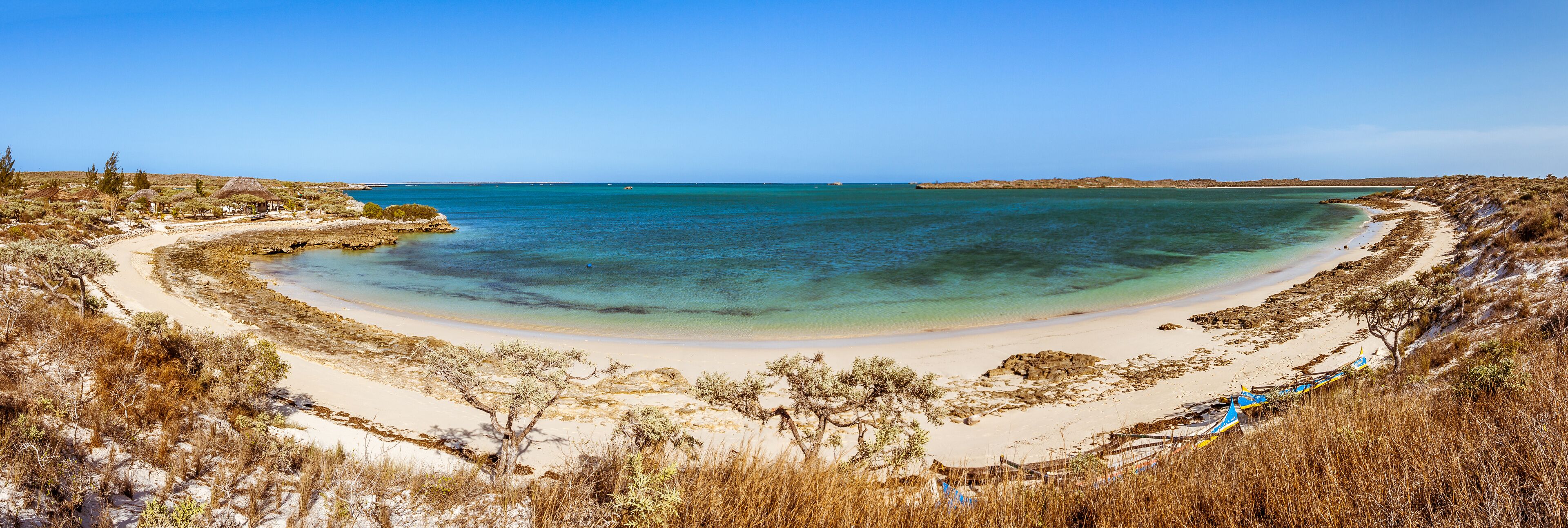 A coastal landscape with a sandy beach, clear turquoise waters, and sparse vegetation.
