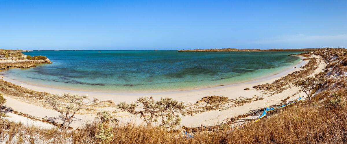 A coastal landscape with a sandy beach, clear turquoise waters, and sparse vegetation.