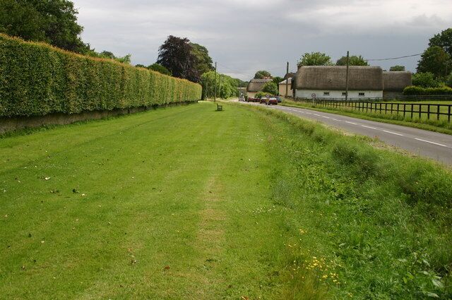 A rural road with a hedge on one side and a building with a thatched roof on the other.