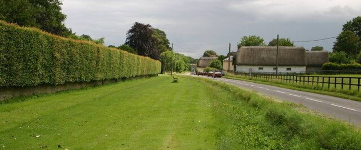 A rural road with a hedge on one side and a building with a thatched roof on the other.