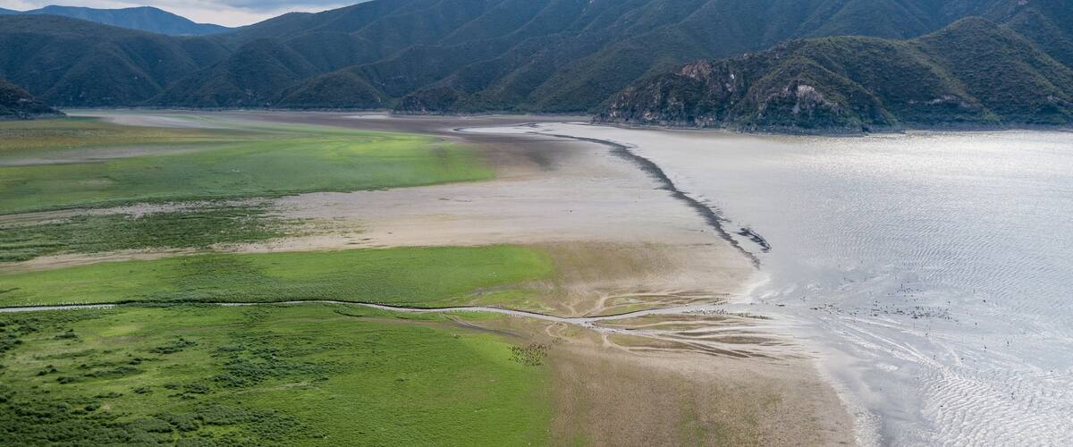 A coastal landscape with a river flowing into the sea, surrounded by green hills and mountains.