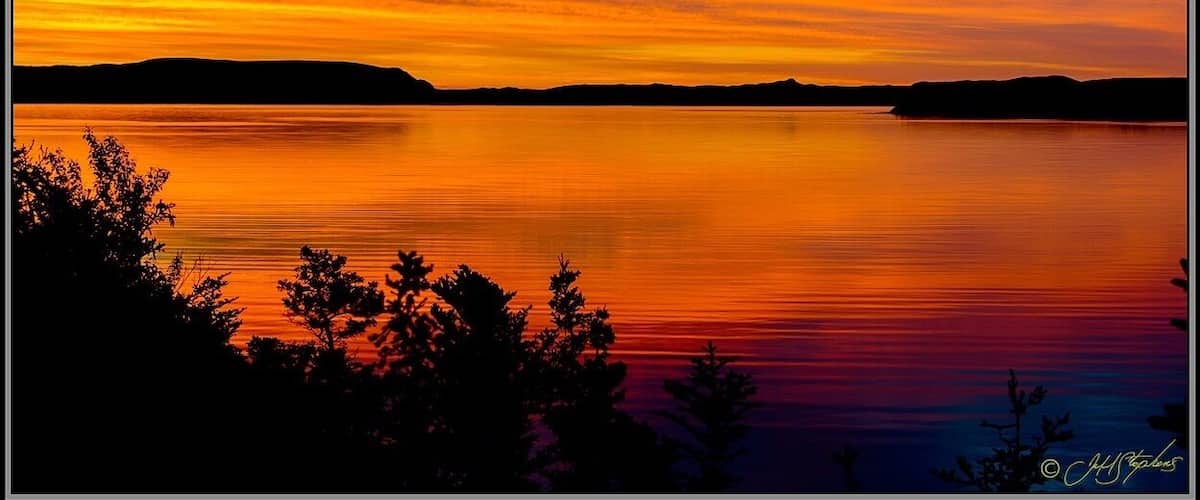 A sunset over a calm lake with silhouetted trees and distant hills.