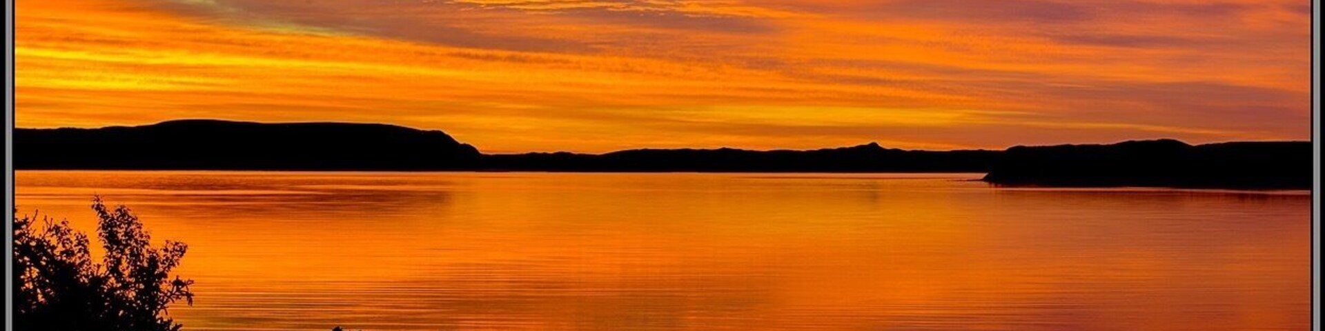 A sunset over a calm lake with silhouetted trees and distant hills.